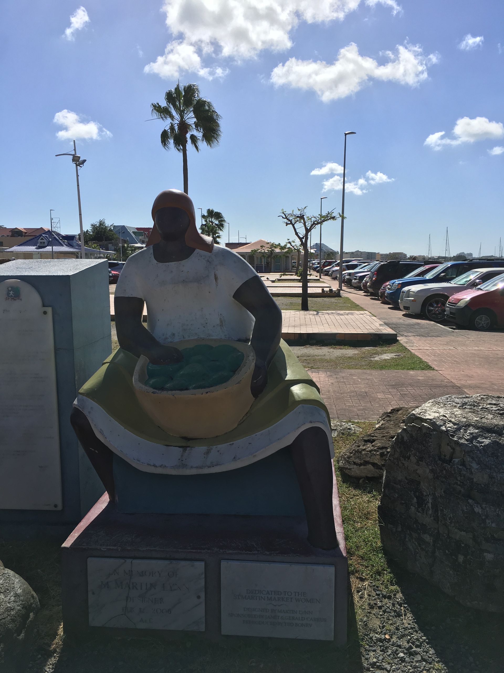 Statue of a woman with a basket, sunny day, outdoor scene with parked cars and buildings in the background.