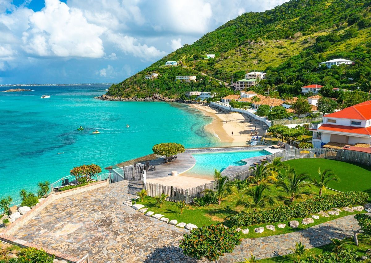 Tropical beach with turquoise water, a white sand beach, and a hillside with buildings and lush greenery.