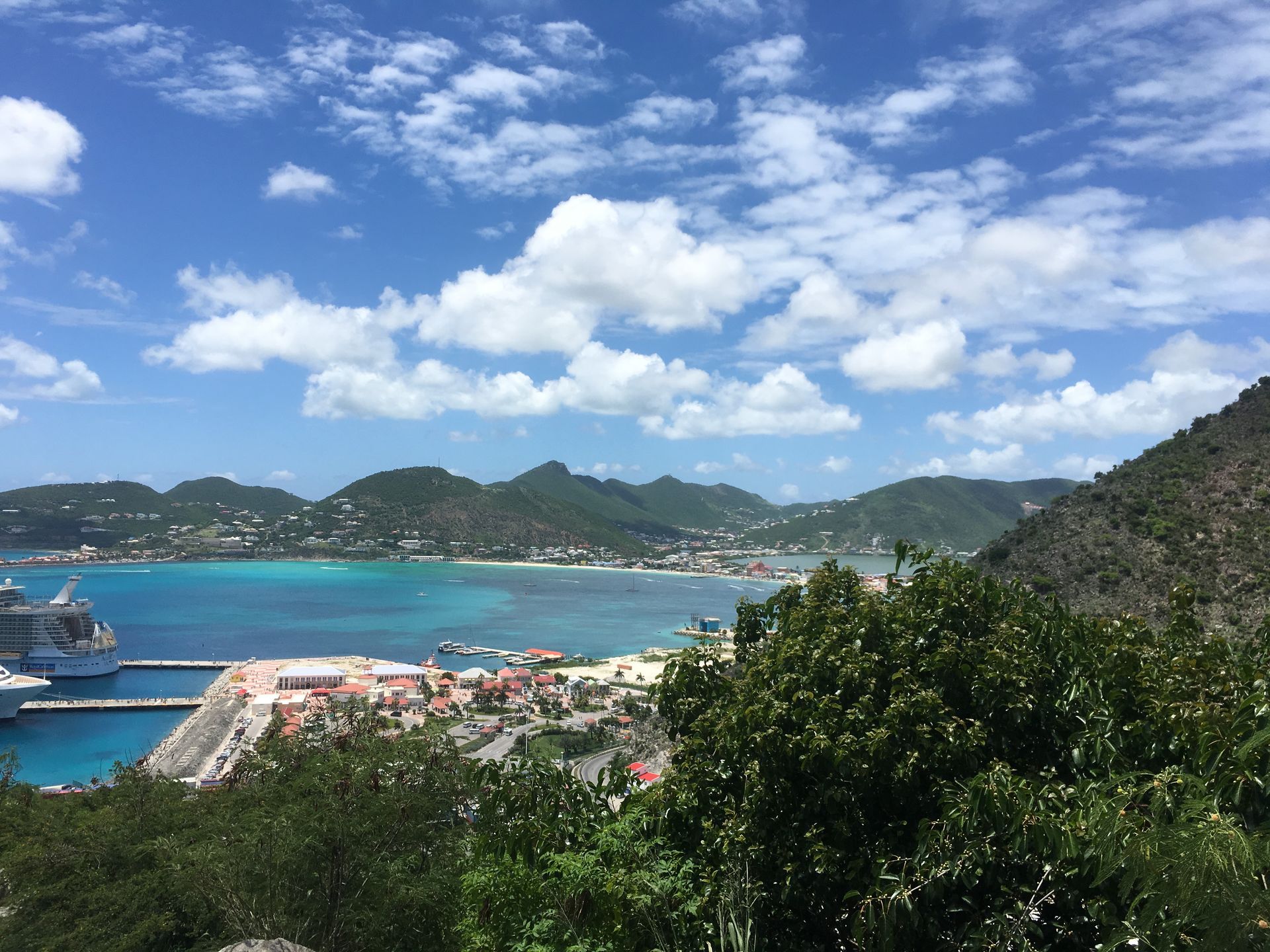 Harbor view: cruise ship, turquoise water, buildings, lush hills under a blue sky with clouds.