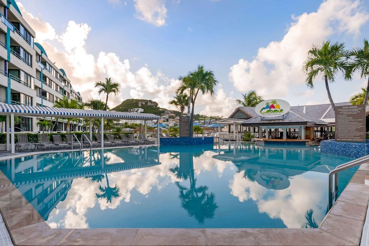 Pool area with a bar, palm trees, and a building, reflecting in the water under a cloudy sky.