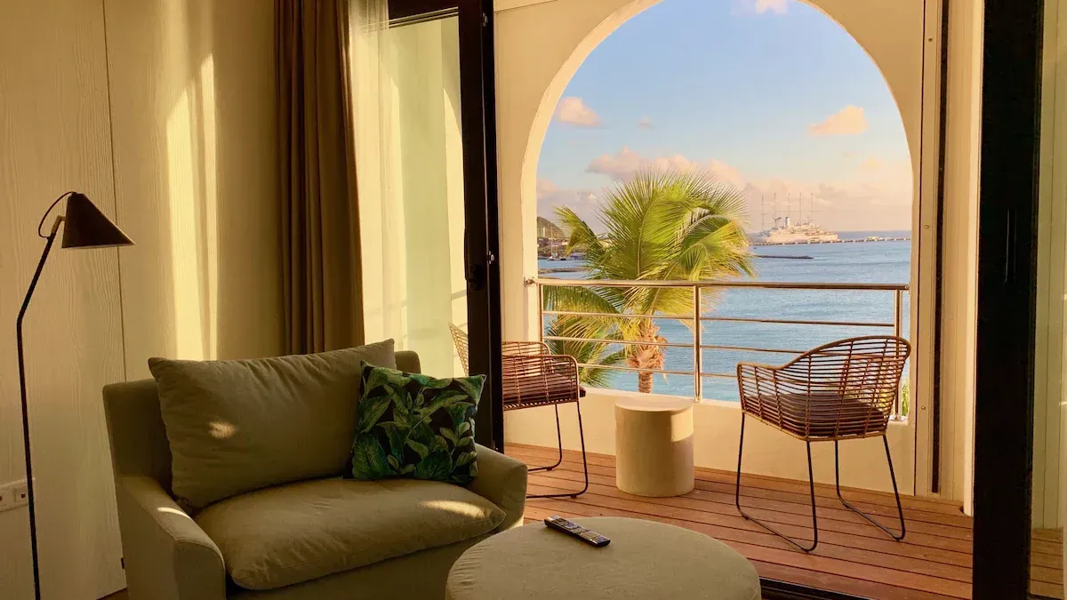 Cozy room with armchair and balcony overlooking the ocean, palm trees, and a ship.