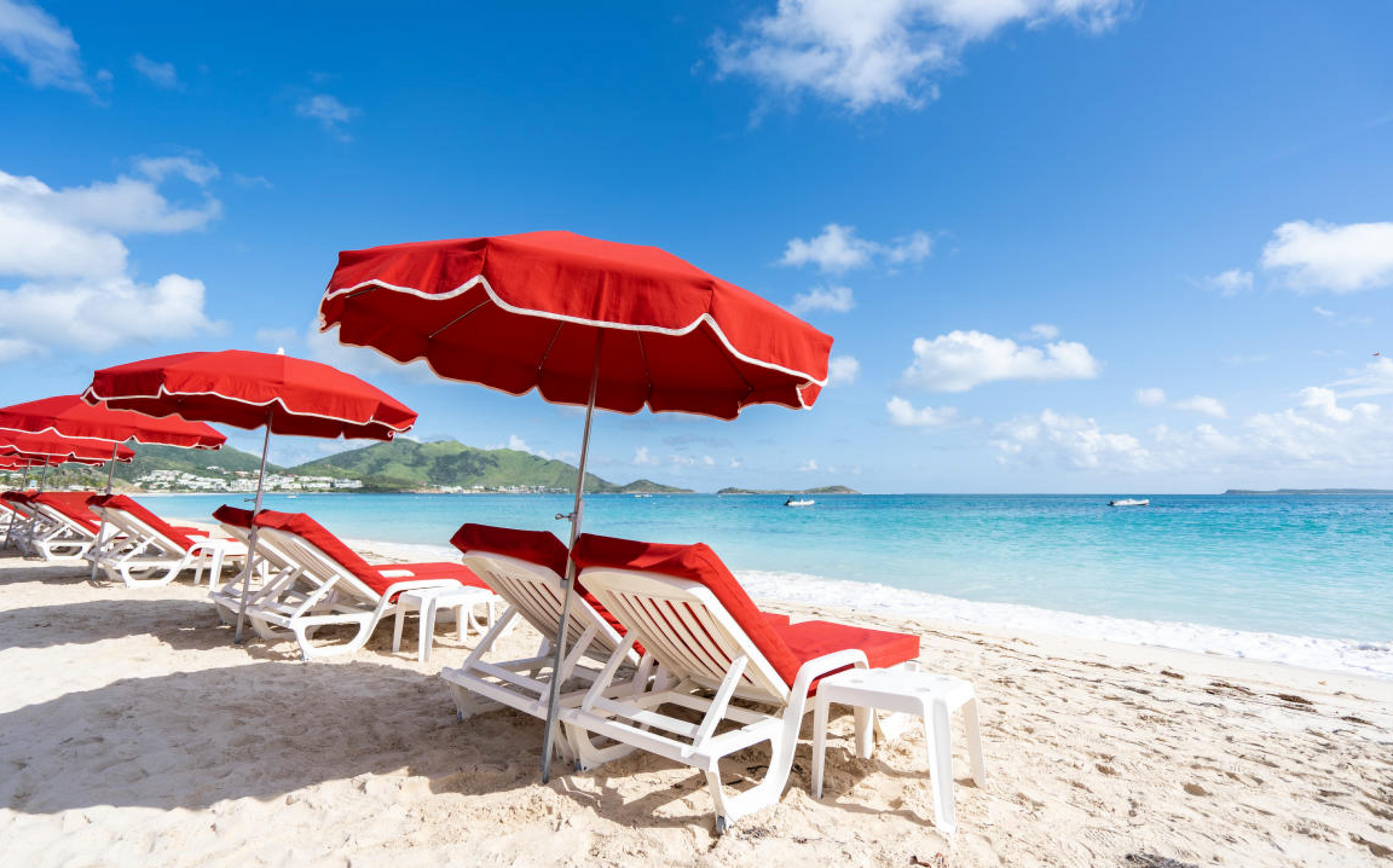 Beach scene with red umbrellas, white lounge chairs, and blue ocean under a sunny sky.