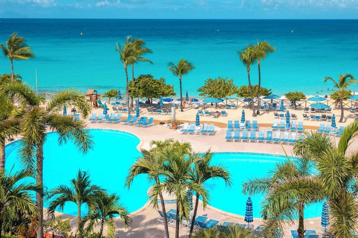 Pool and beach scene with turquoise water, palm trees, and blue lounge chairs.