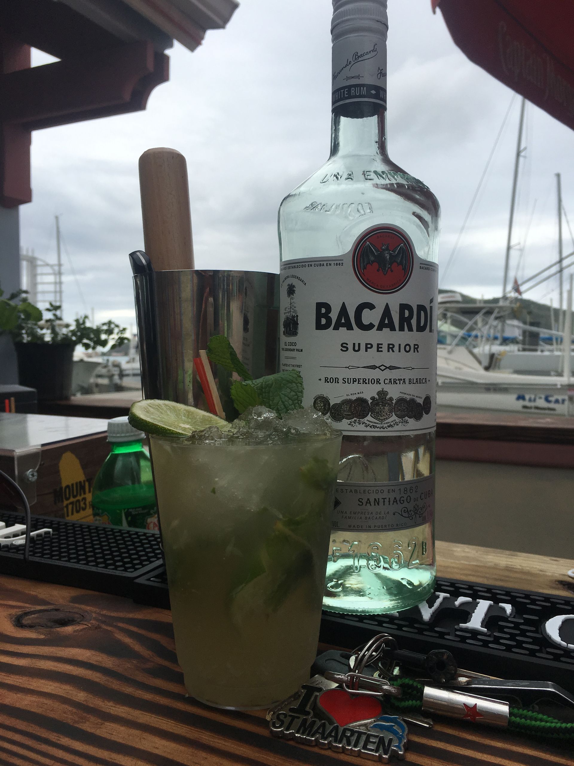 Mojito cocktail in a glass, with Bacardi rum bottle behind it. On a wooden bar, harbor background.