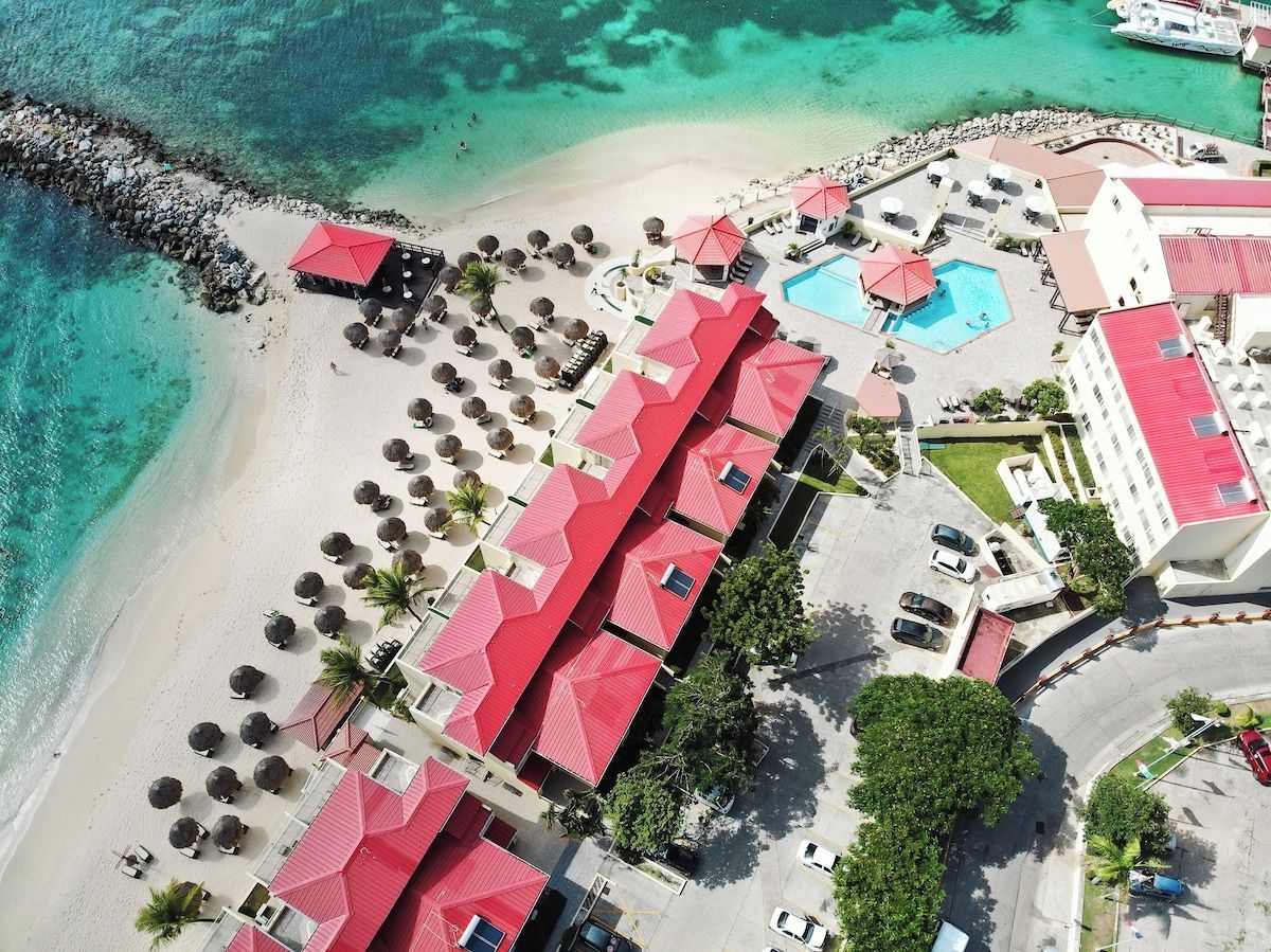 Aerial view: Red-roofed resort, beach, turquoise water, small pool, palm trees, and clear sky.