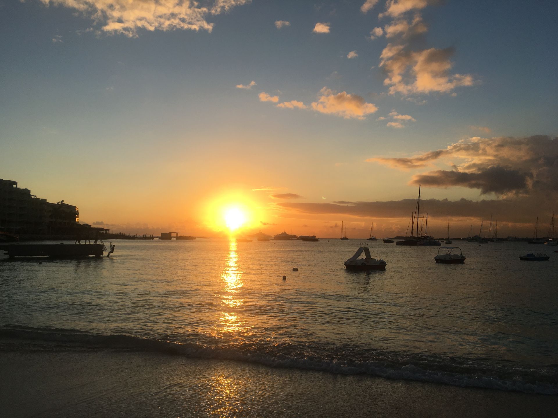 Sunset over calm water, with boats and a few clouds in the sky, near a beach.