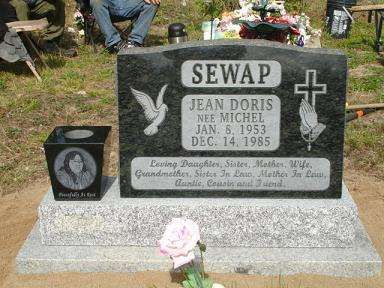 Gravestone with angel and cross carvings, white plaque, and a small memorial vase in a cemetery.