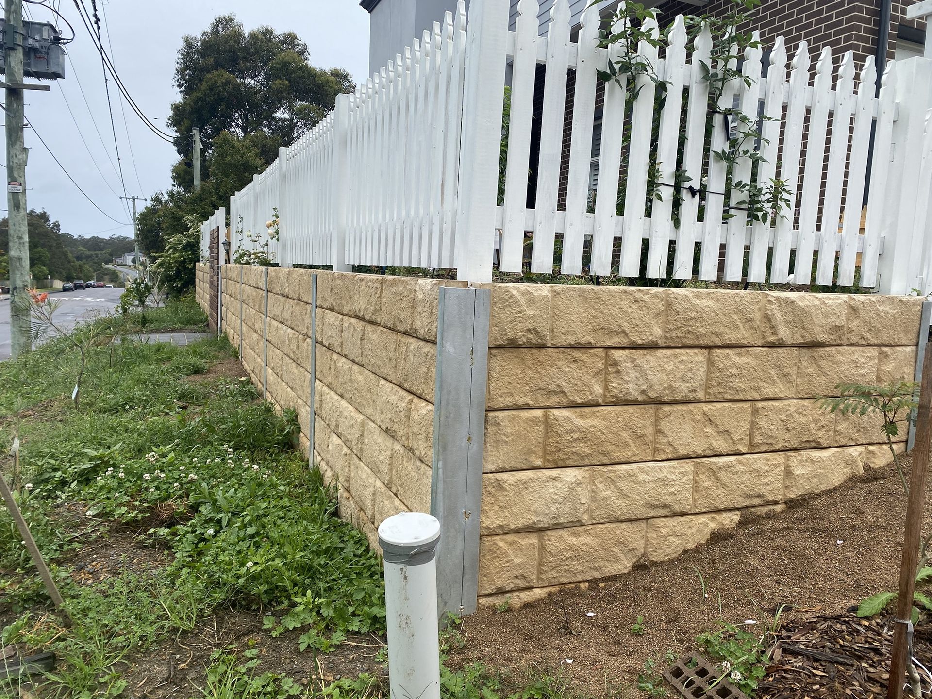 A Retaining Wall Made of Tan Blocks Supports a White Picket Fence — Concrete Sleepers NSW - Port Macquarie in Newcastle, NSW