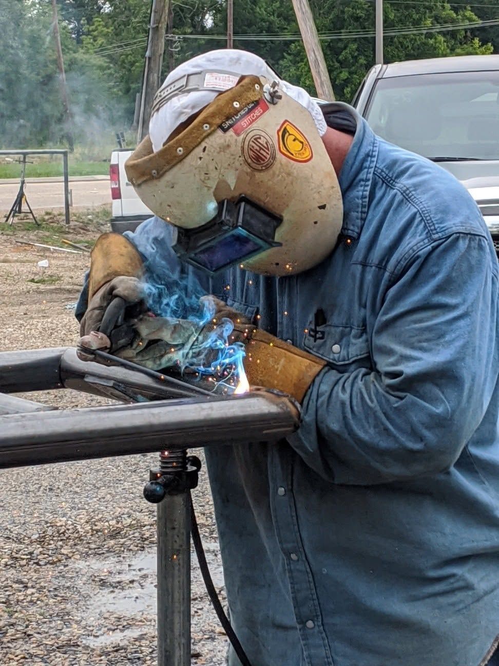 Welder in blue jacket and helmet welding a metal frame outdoors, with smoke and sparks visible
