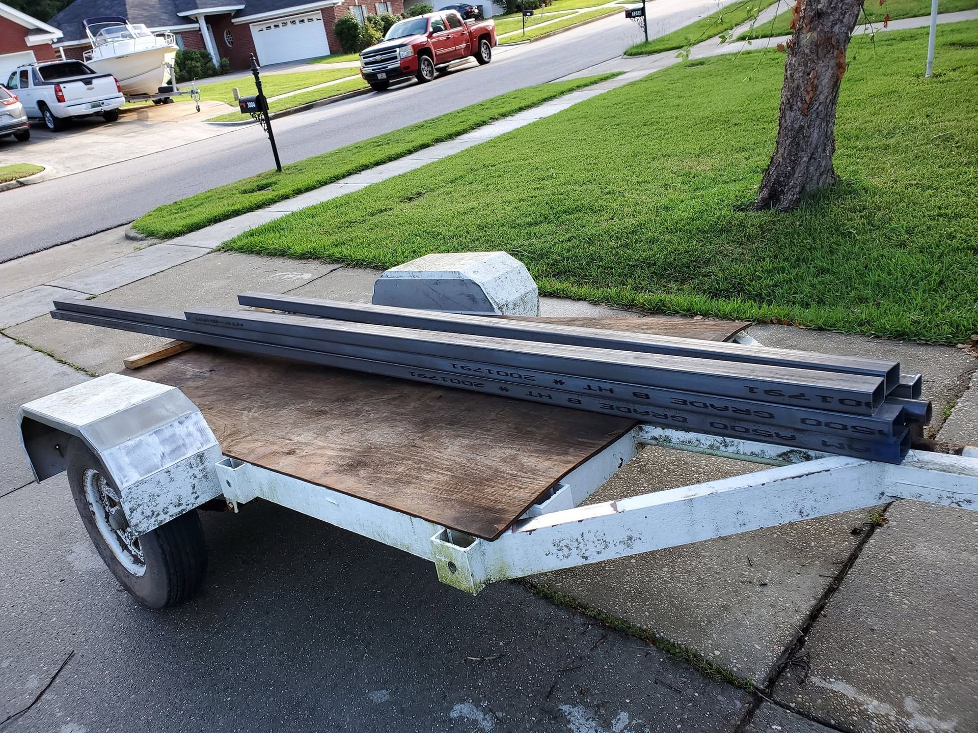 Utility trailer carrying long metal beams on a driveway near a suburban lawn.