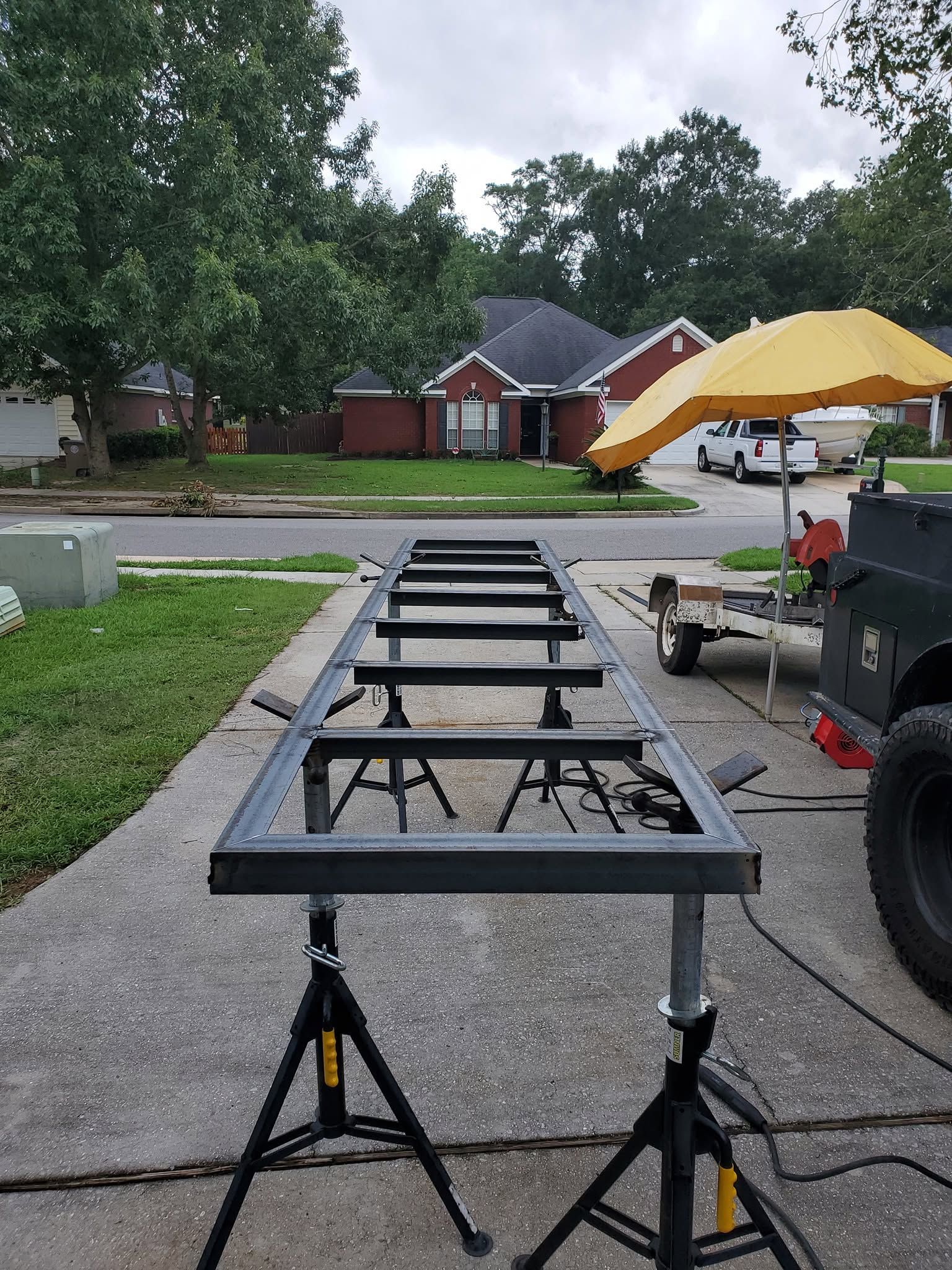 Black metal trailer frame on jack stands in a driveway, with a yellow umbrella and truck nearby.