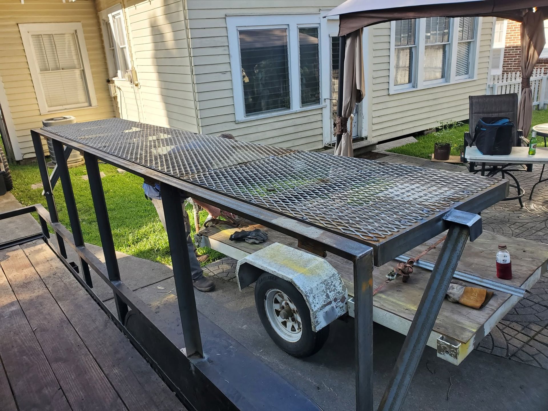Metal trailer with mesh ramp beside a house deck, parked in a driveway.