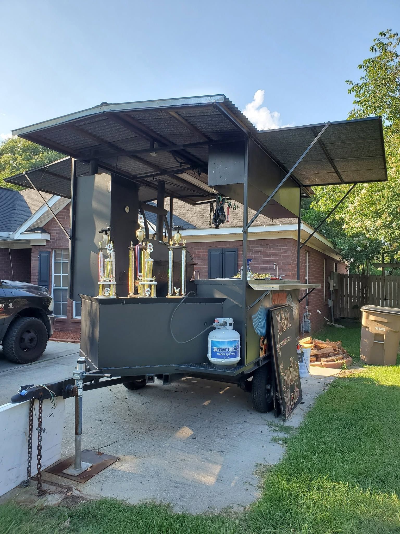 Outdoor pizza oven trailer with black canopy, propane tanks, and a blue gas cylinder in a driveway.