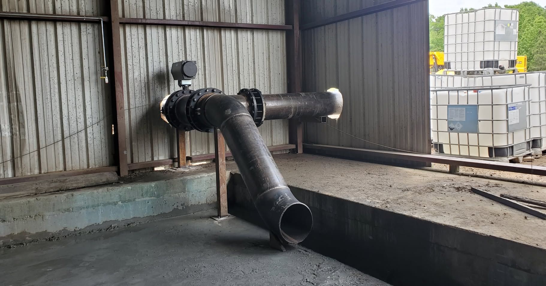 Black industrial pipe jutting through a warehouse wall into a concrete pit.