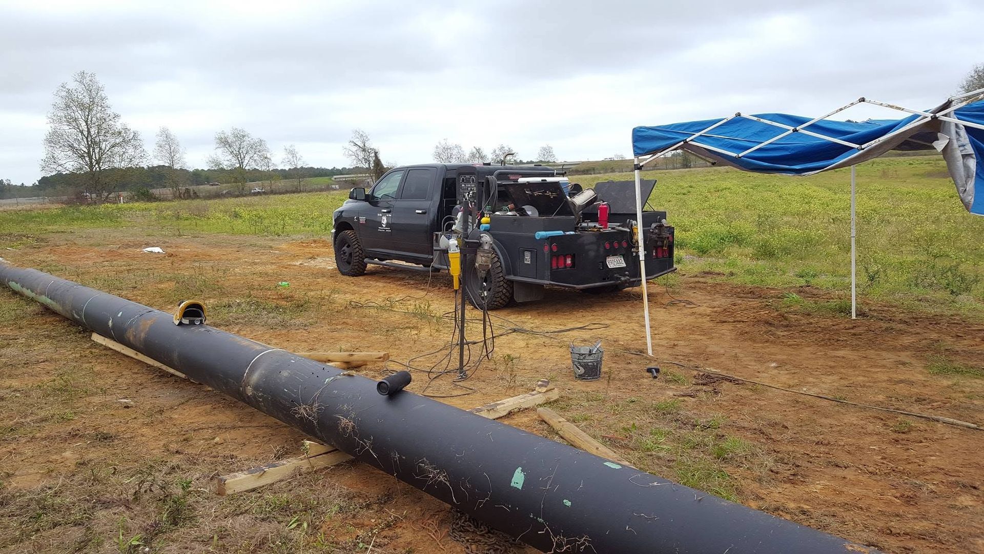 Black pipeline in a muddy field with a pickup truck and blue tarps near a fence
