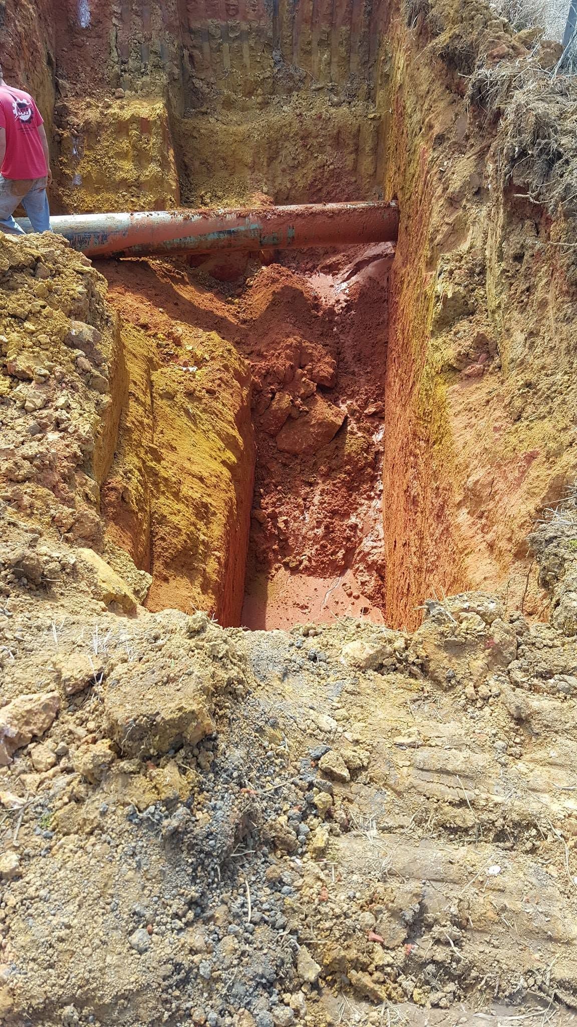 A muddy excavation pit with a large pipe or log spanning the top and a person partially visible at left.
