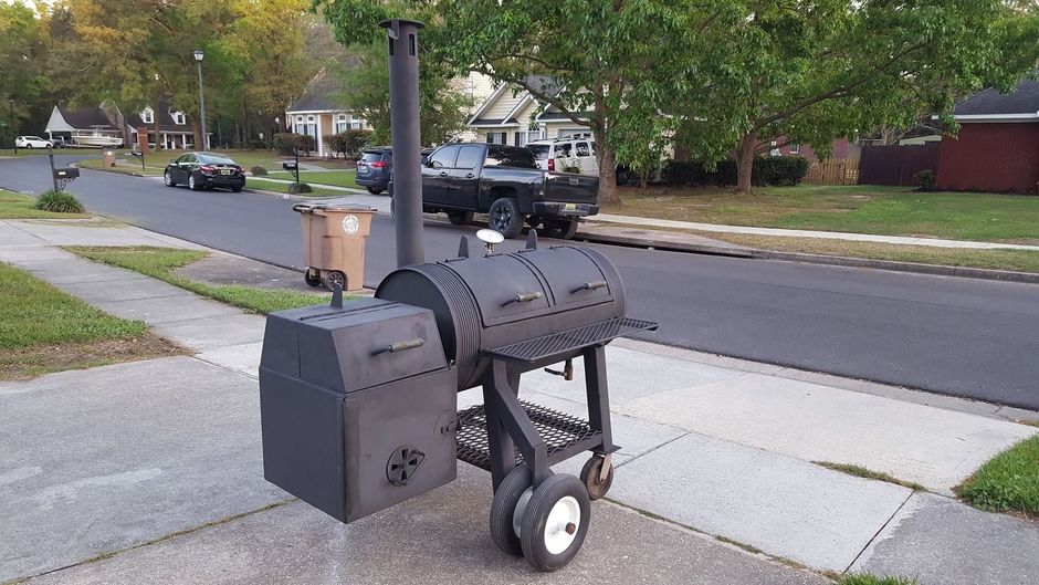Black barrel-style smoker grill on a wheeled stand beside a suburban street and sidewalk.