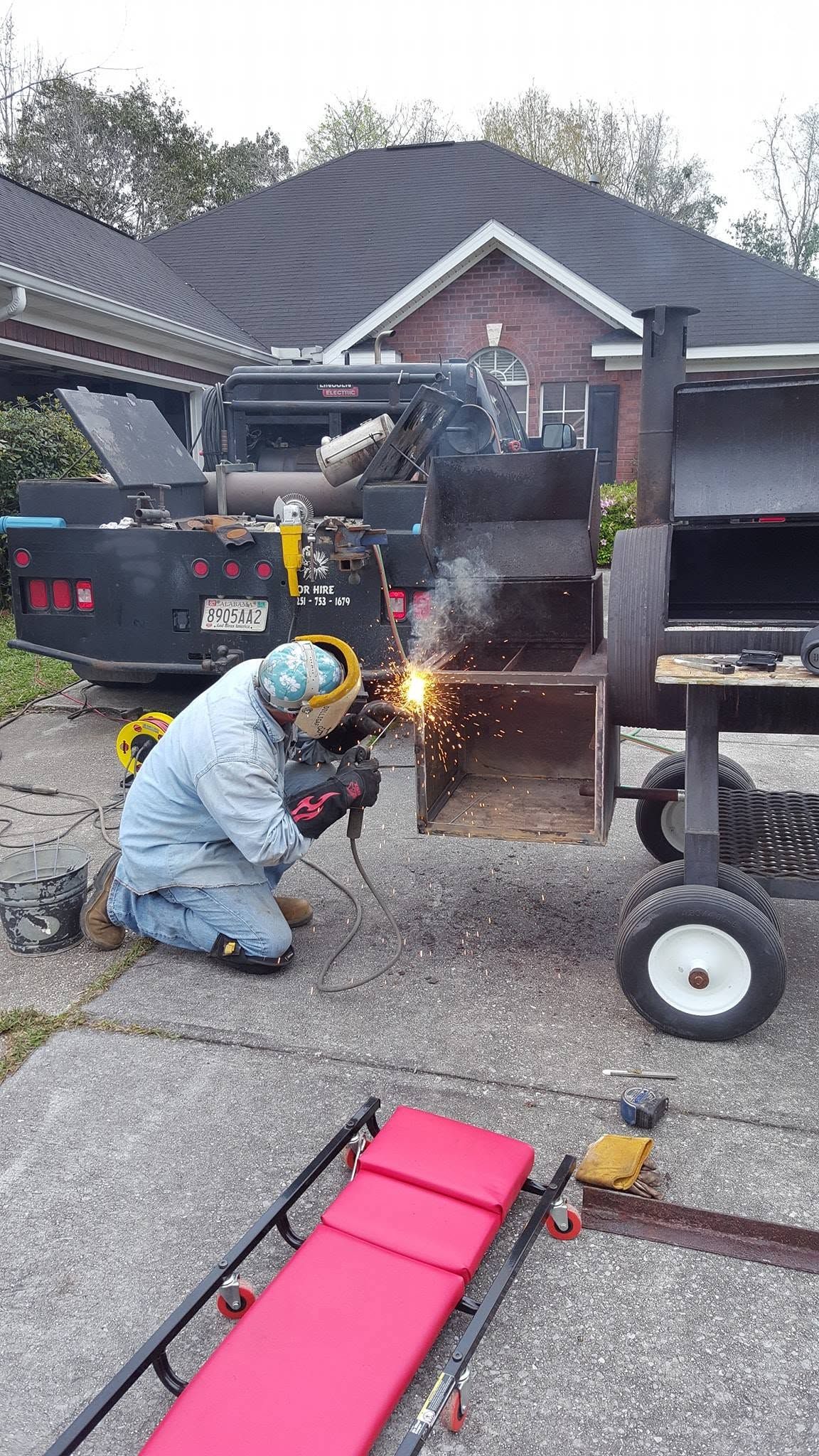 Person welding metal on a trailer outdoors, with sparks flying beside a driveway.