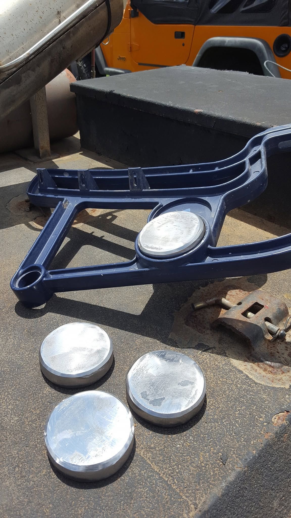 Blue metal machine part with three silver disks on pavement near an orange machine