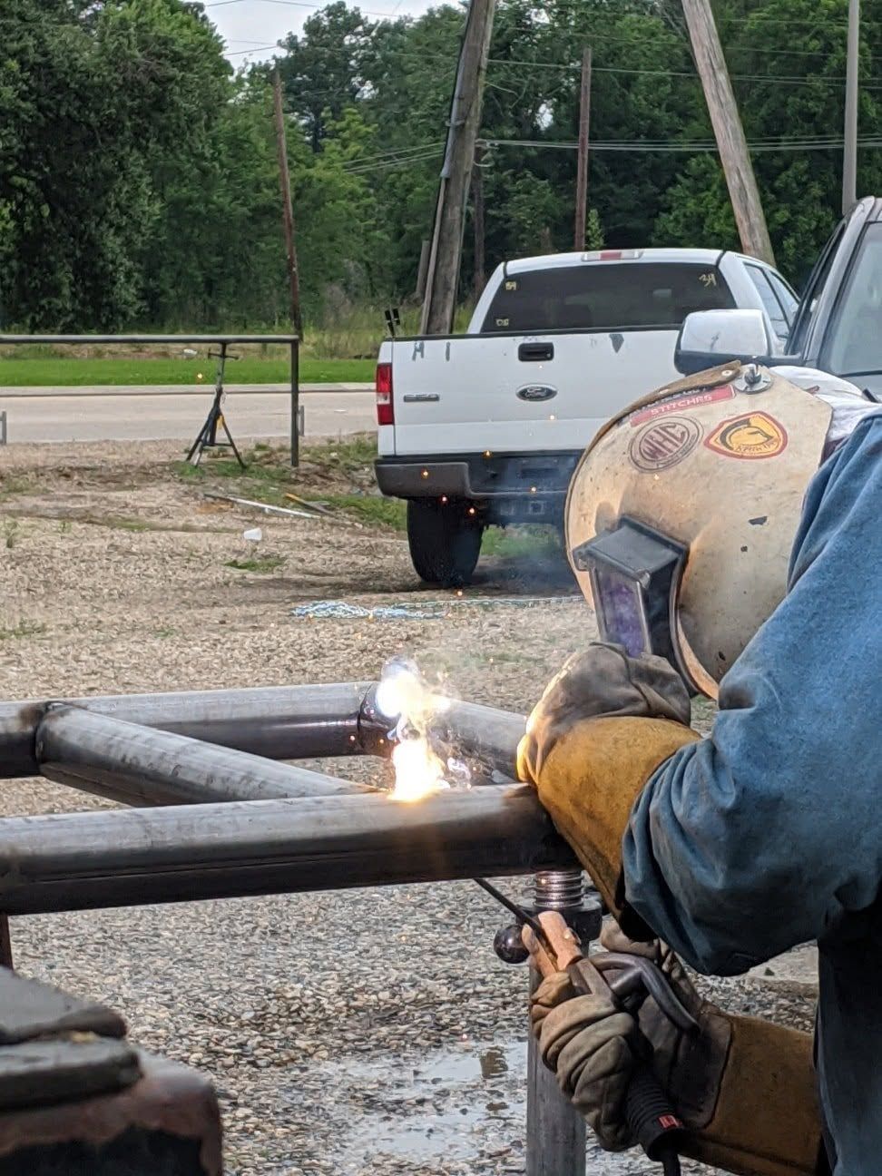 Welder in protective gear welding metal pipes outdoors near a white pickup truck.