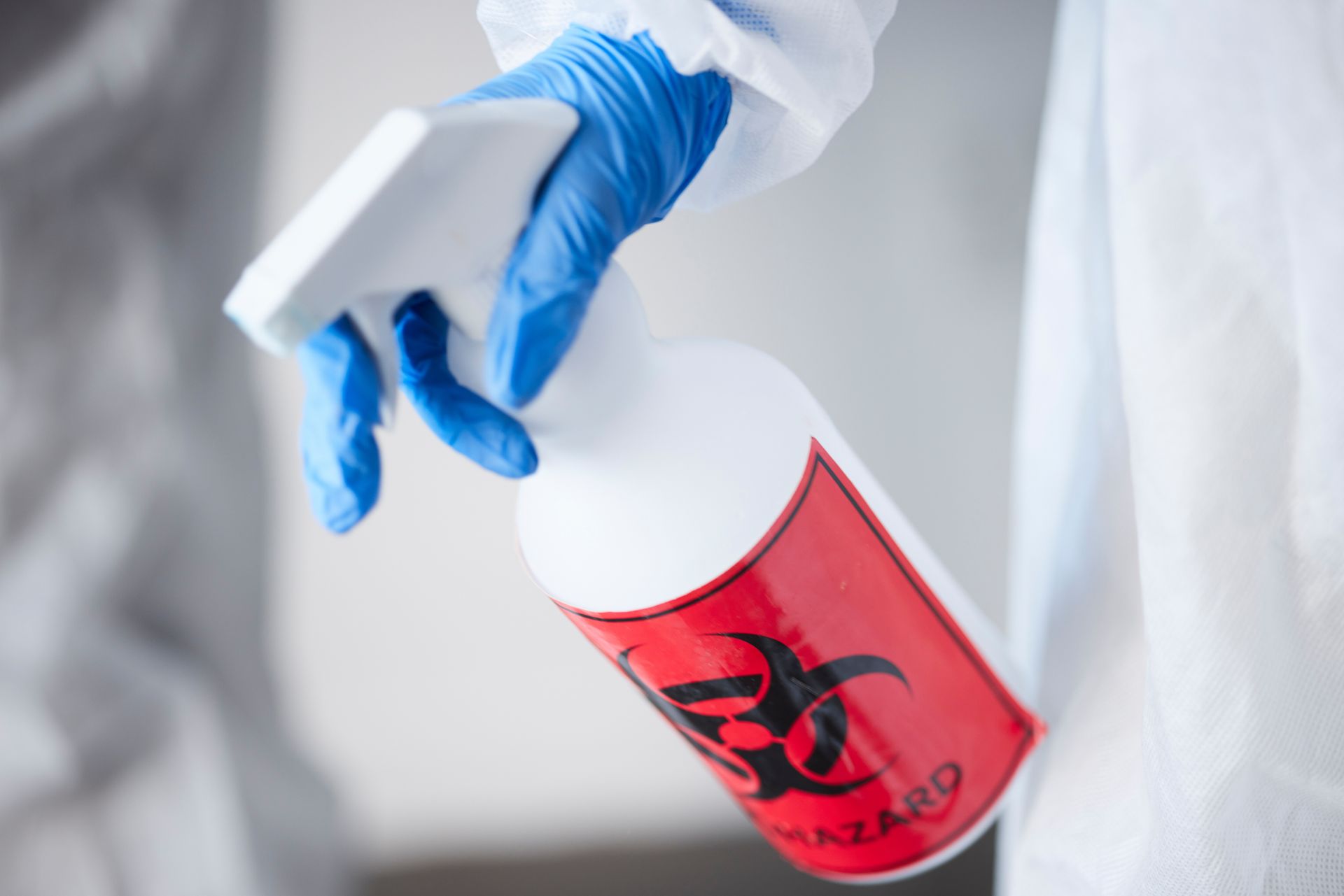 A person wearing blue gloves is holding a spray bottle with a biohazard label.