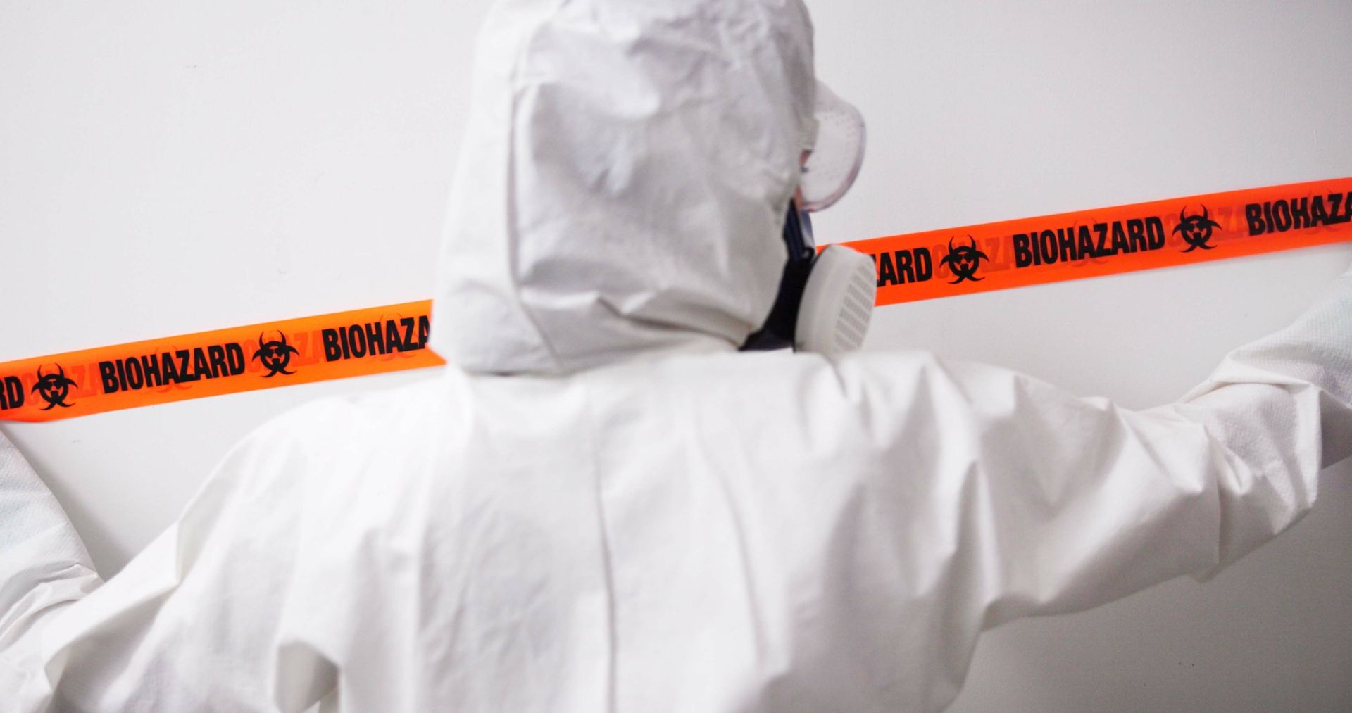 A person in a protective suit is standing in front of a wall with a biohazard tape.