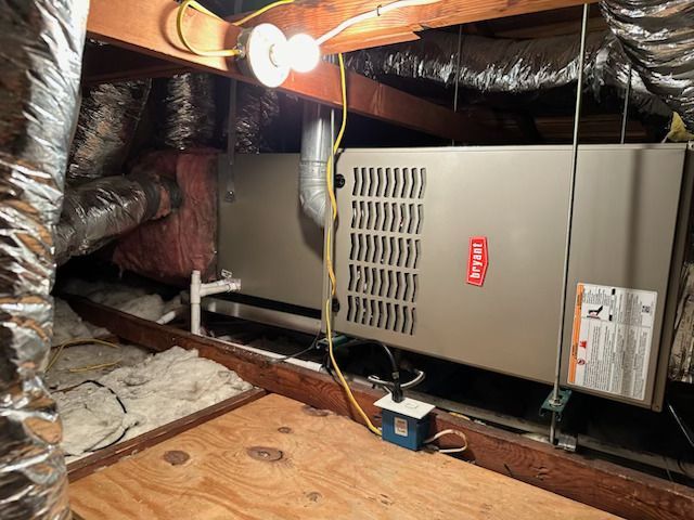 Attic HVAC unit with exposed ductwork, light bulb, and insulation under a sloped roof.