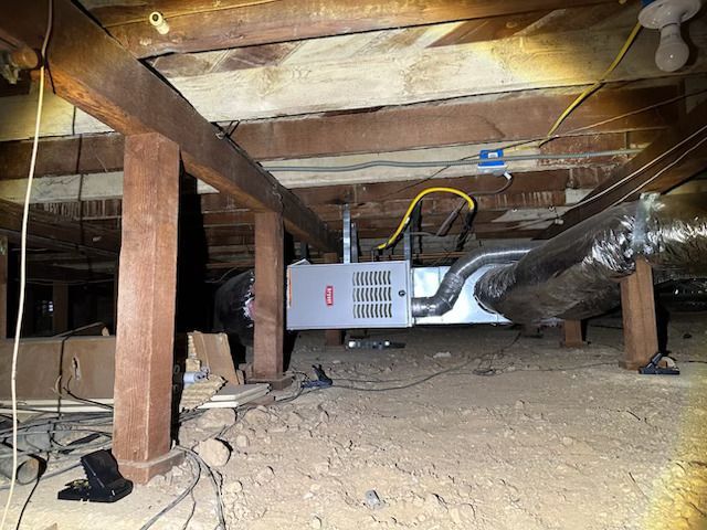 Crawlspace with exposed wooden joists, a duct, and a small HVAC unit amid dirt and debris.