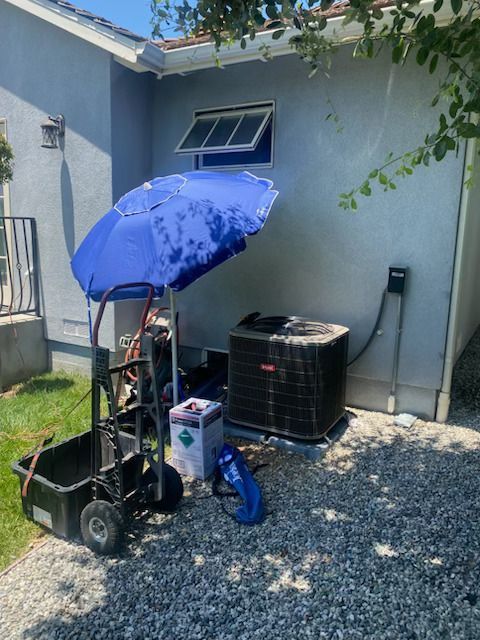 Blue umbrella-covered cart and equipment beside a house’s exterior wall on a gravel yard.
