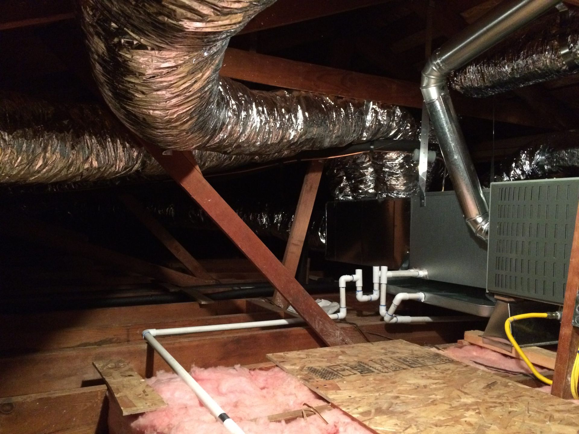 Attic with exposed ductwork, pipes, insulation, and a plywood floor opening illuminated by a flashlight