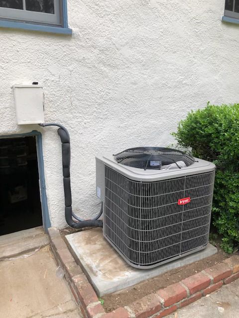 Outdoor air conditioner unit beside a stucco wall, with conduit and a bush nearby.