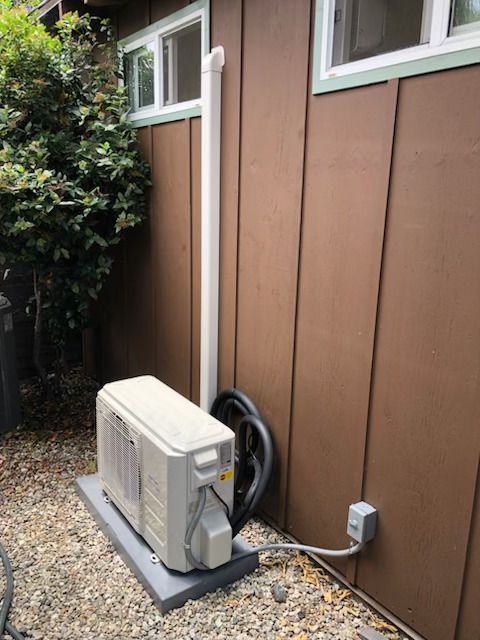 Outdoor air conditioner unit beside a brown house wall with a coiled hose and gravel ground