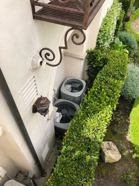 Two outdoor HVAC units beside a house, bordered by a neatly trimmed hedge and wall-mounted lights.