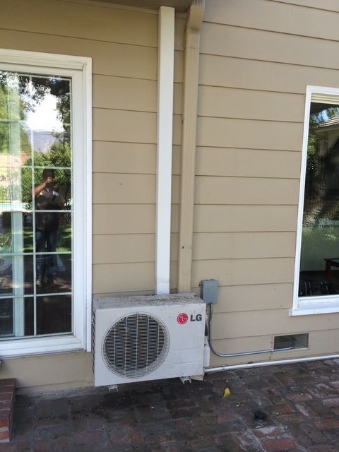 LG outdoor AC unit mounted beside a beige house wall between two windows.