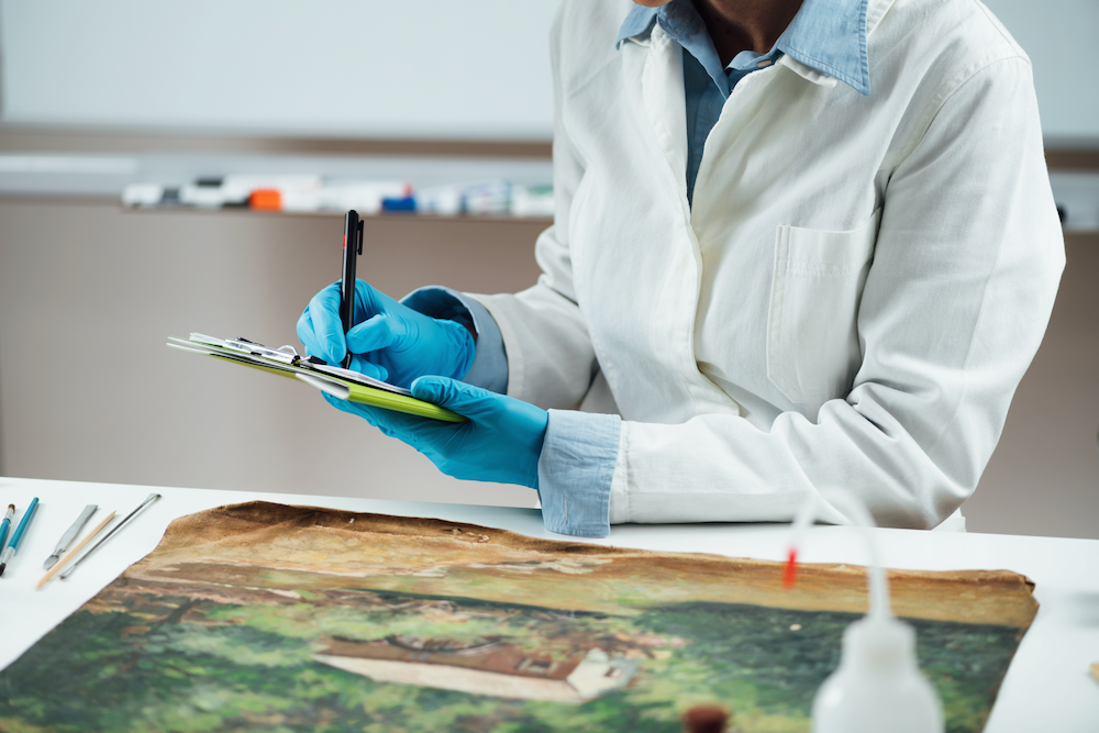 A man in a lab coat and blue gloves is writing on a clipboard over a painting.