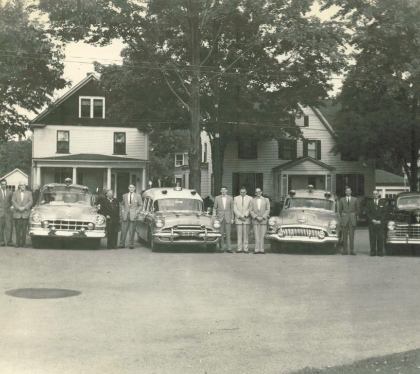 Group of men standing with vintage emergency vehicles in front of two-story houses.