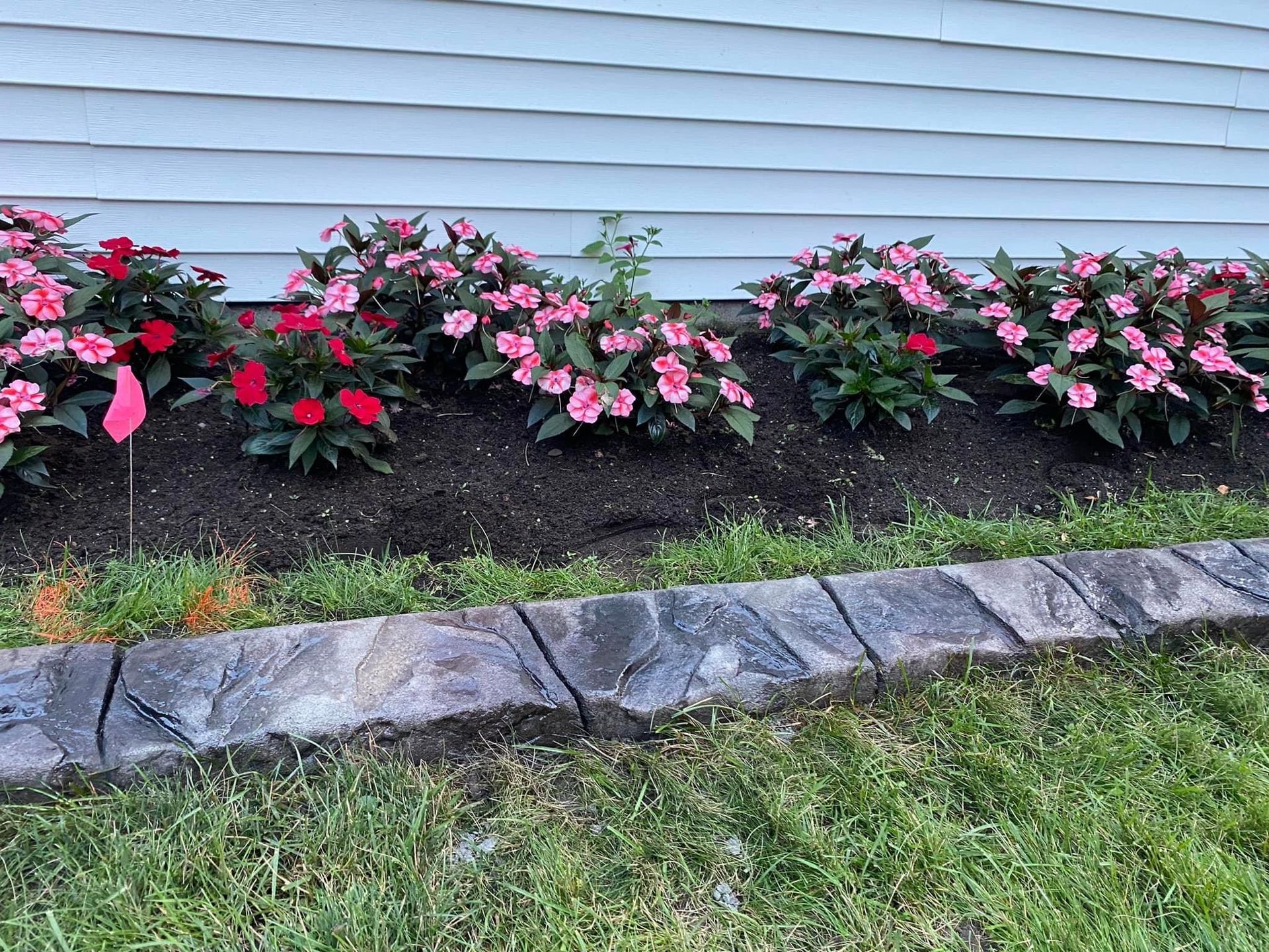 Pink and red flowers in a landscaped garden bed beside a house, bordered by stone edging