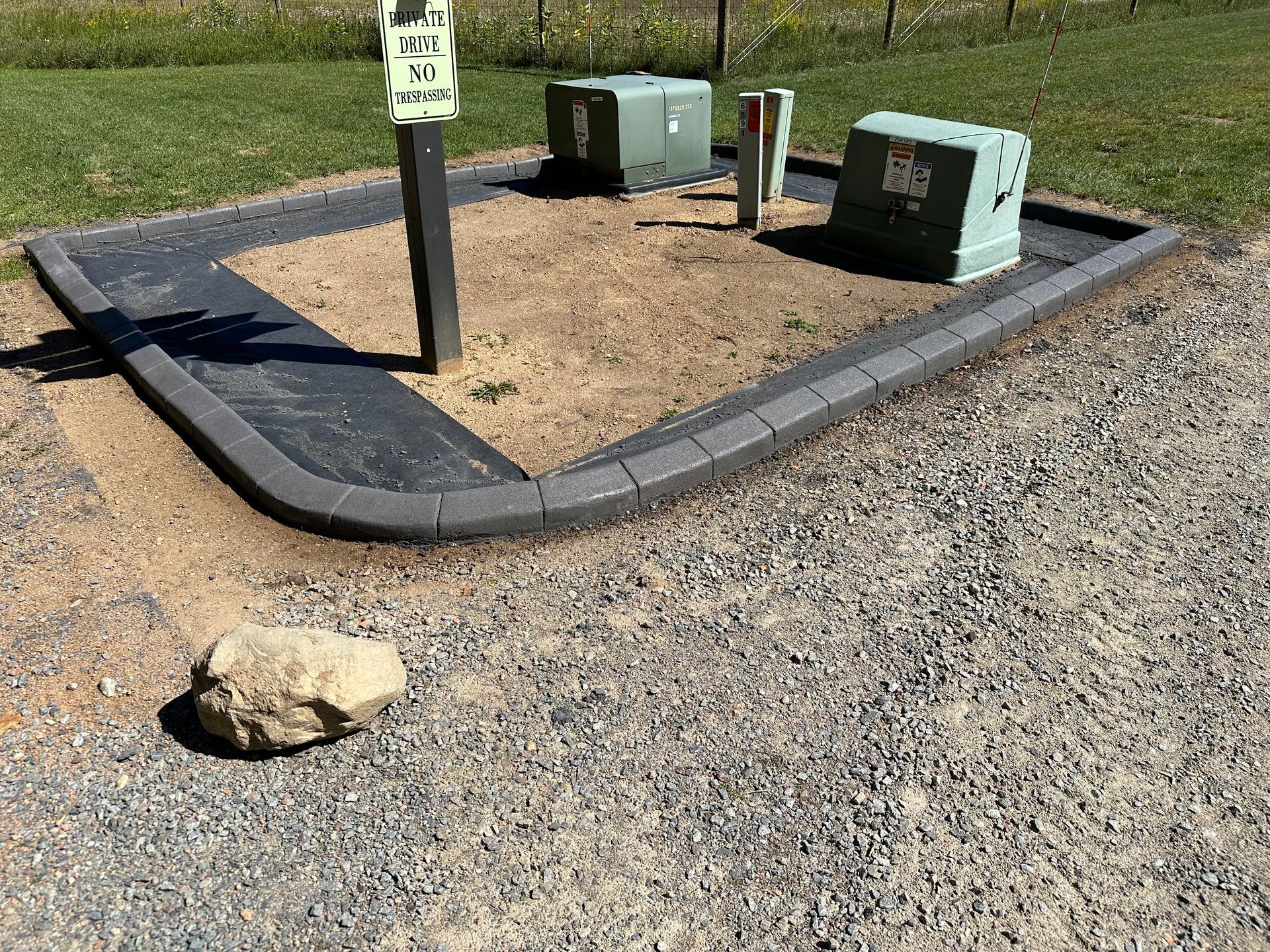 Small utility enclosure with green electrical boxes on gravel beside a parking lot, with a signpost and rocks.