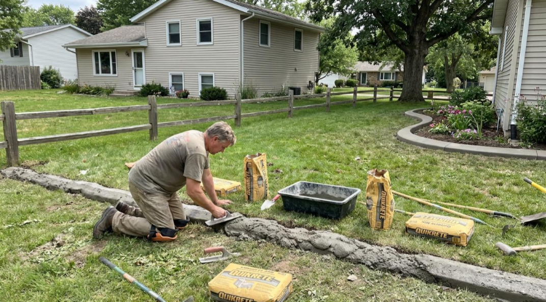 Person kneeling on grass beside fence posts in a yard, working with a tool near a house and tree.