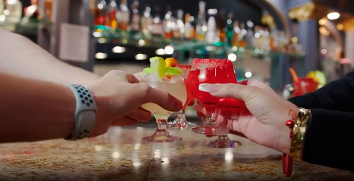 Hands clinking colorful cocktails together at a bar counter, with blurred bottles in the background