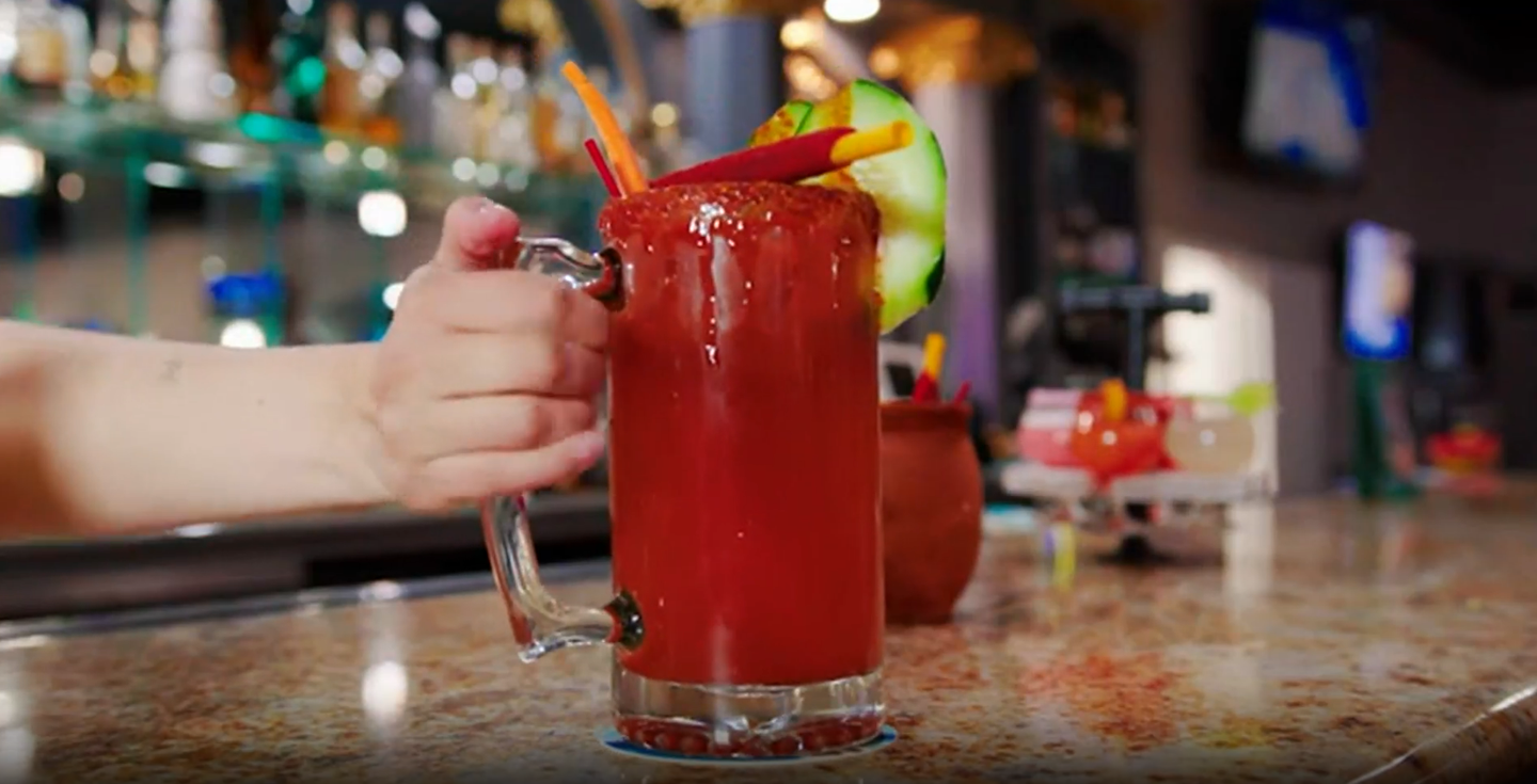A tall red cocktail with a lemon garnish on a bar counter, held by a hand