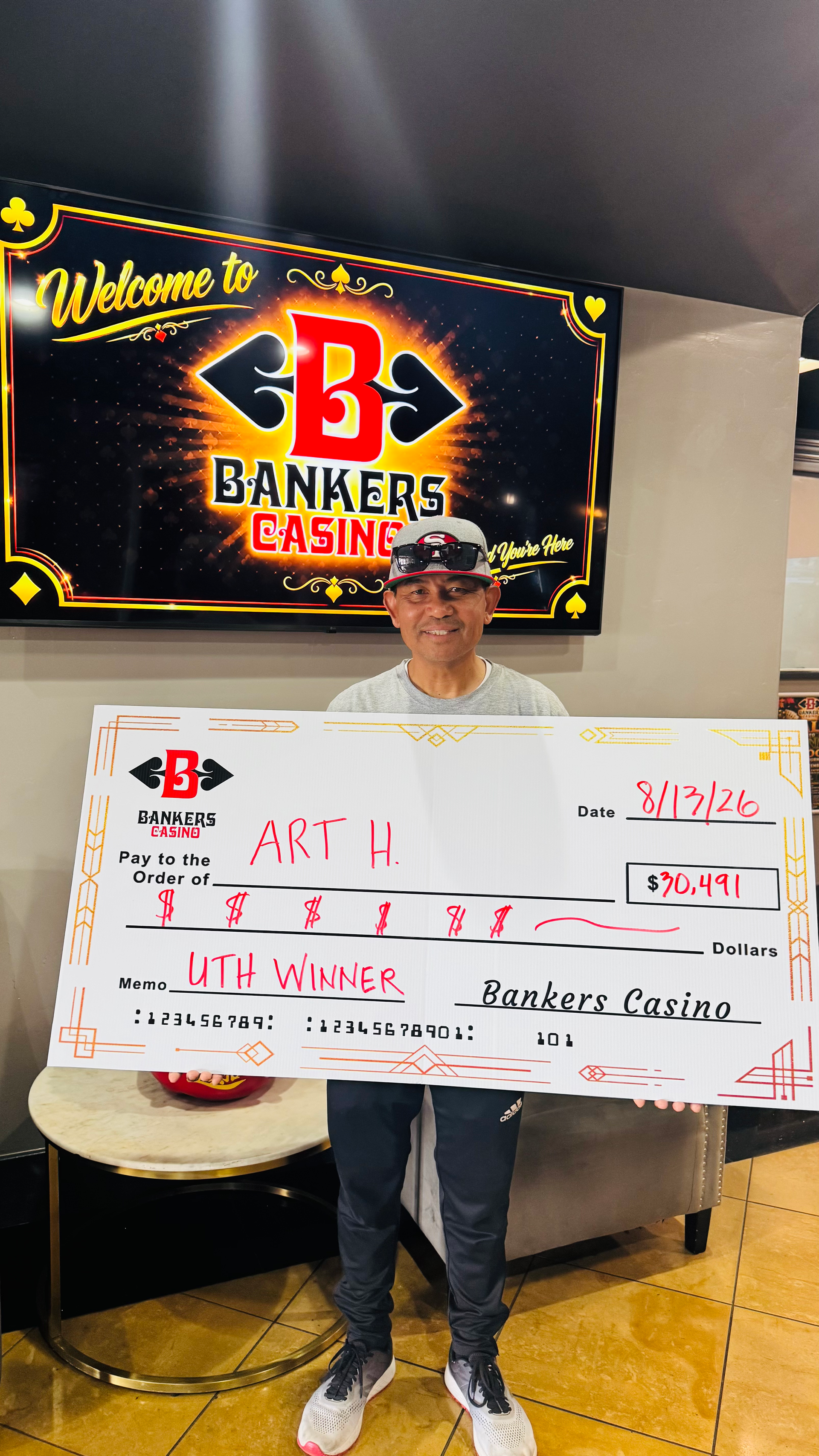 Man holding a large white check in front of a “Bakers” sign, indoors