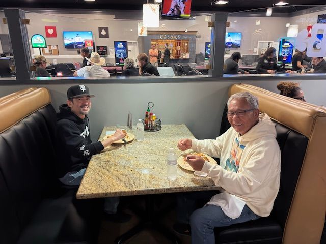 Two people eating at a restaurant booth with a polished table and busy dining area behind them