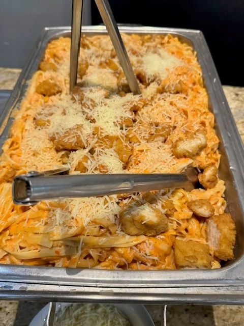 Tray of baked pasta or casserole topped with grated cheese, served with metal tongs in a buffet line