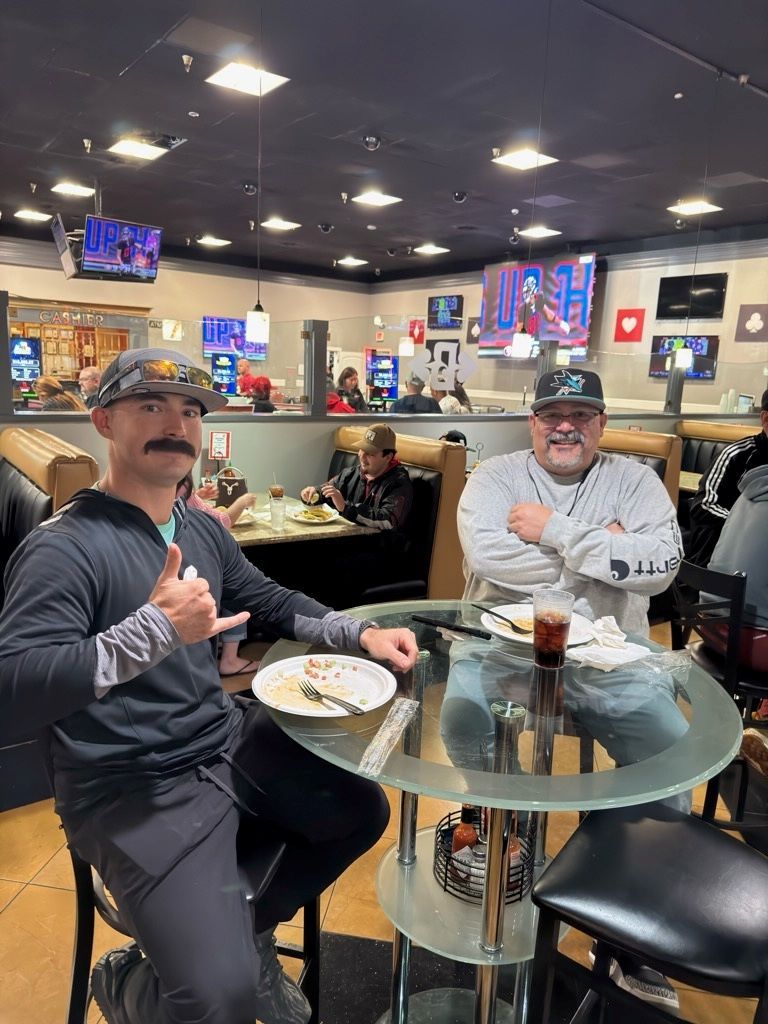 Two men sit at a restaurant table, one holding a drink and smiling in a casual diner setting.