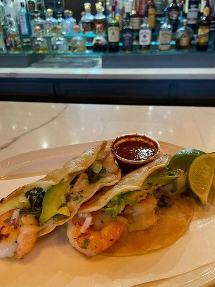 Two tacos on a plate with salsa and lime wedges, at a bar with liquor bottles in the background