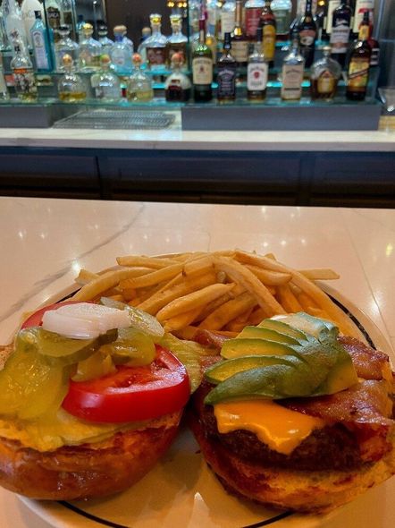 Two burgers with pickles and cheese beside fries on a counter, with a bar of liquor bottles behind them