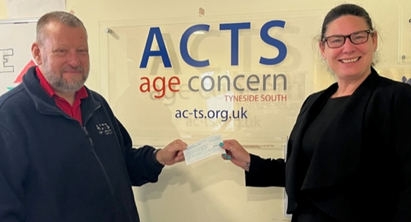 Man in blue jacket hands a check to woman in black jacket in front of an
