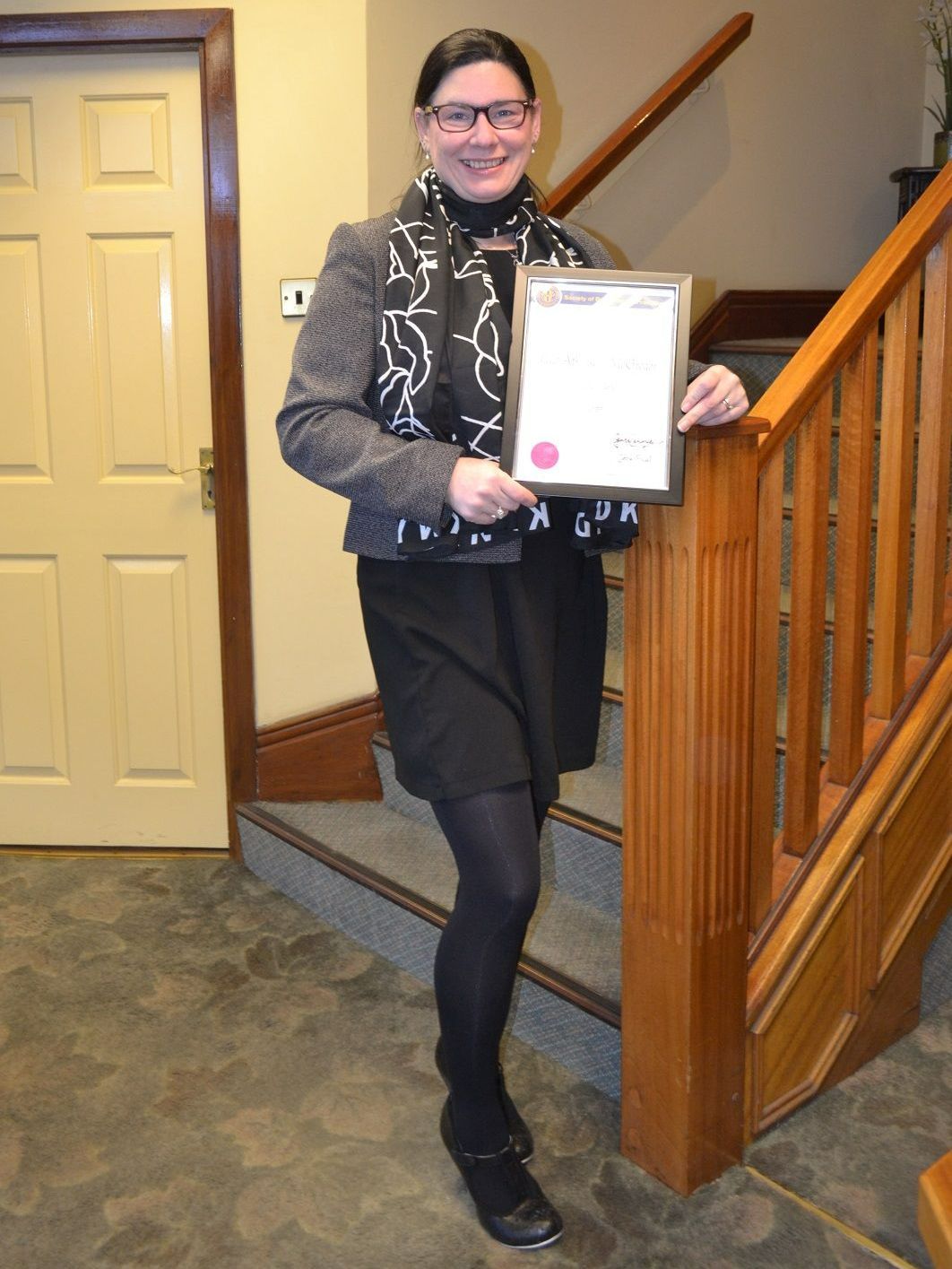 Woman in black outfit holding a framed certificate, smiling on wooden staircase.