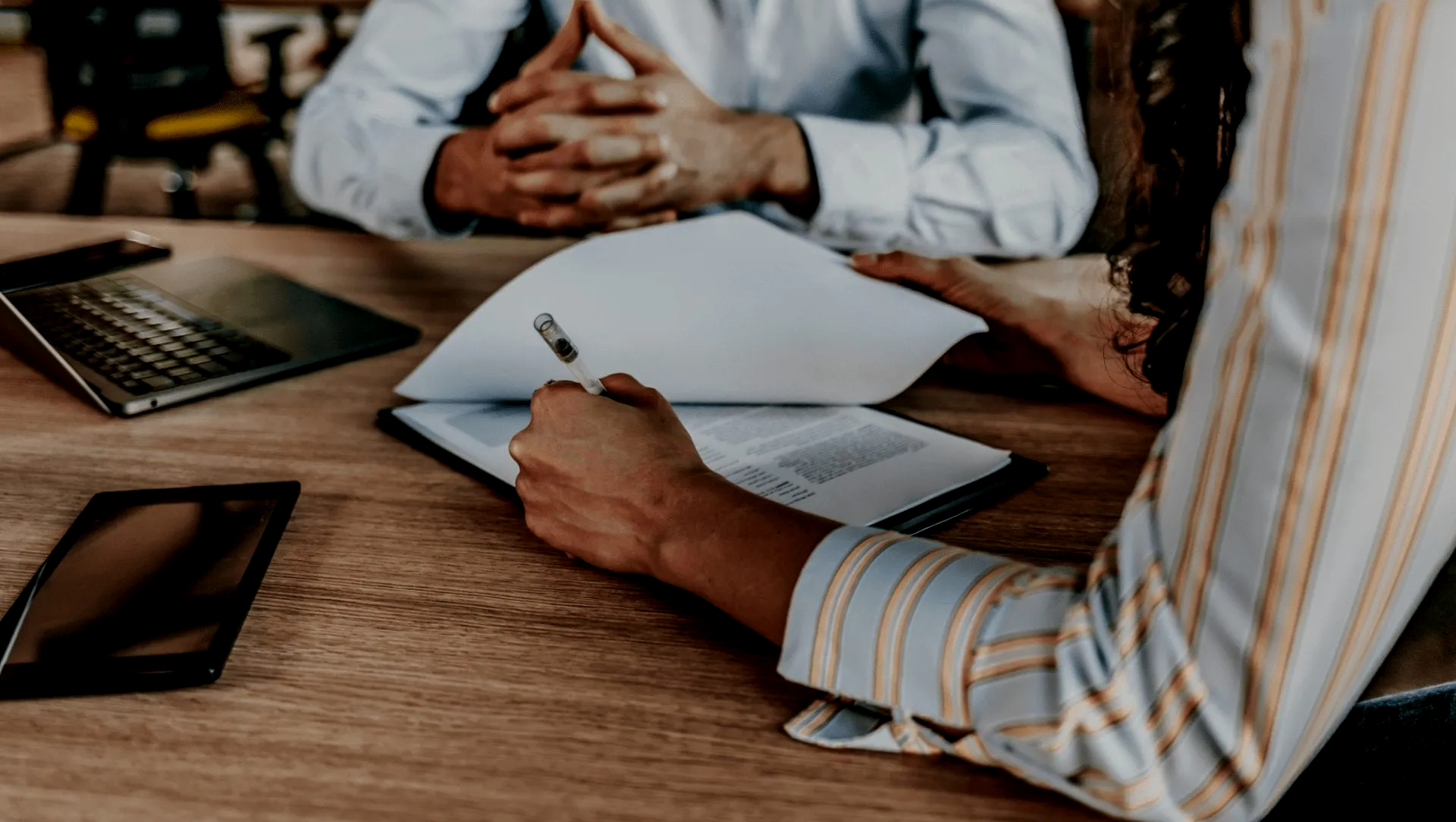 Person signing document at table with another person, laptop, and tablet.